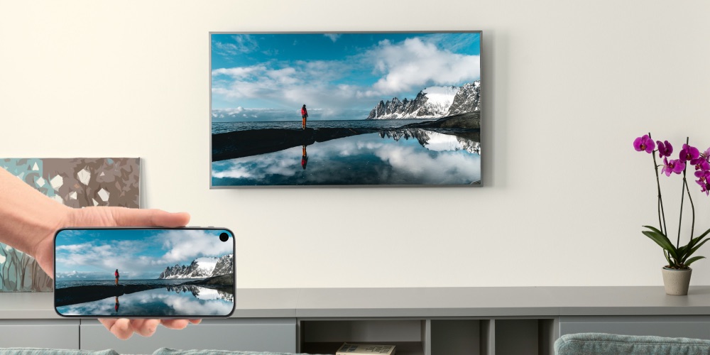 A hand holding an iPhone that is casting an image of a person in a landscape of mountains and water to a wall-mounted Samsung TV